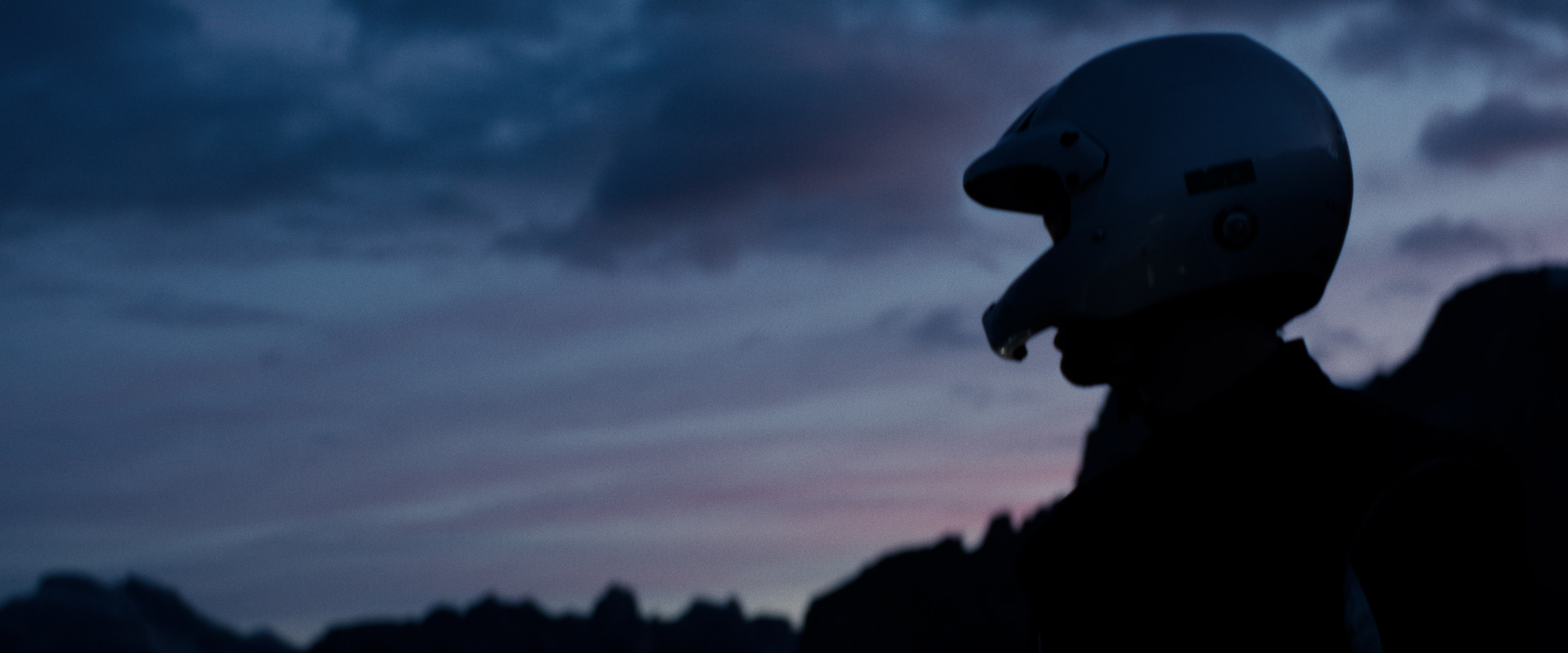 Silhouette of a helmeted racing driver at dusk, mountain skyline faintly visible under violet sky.