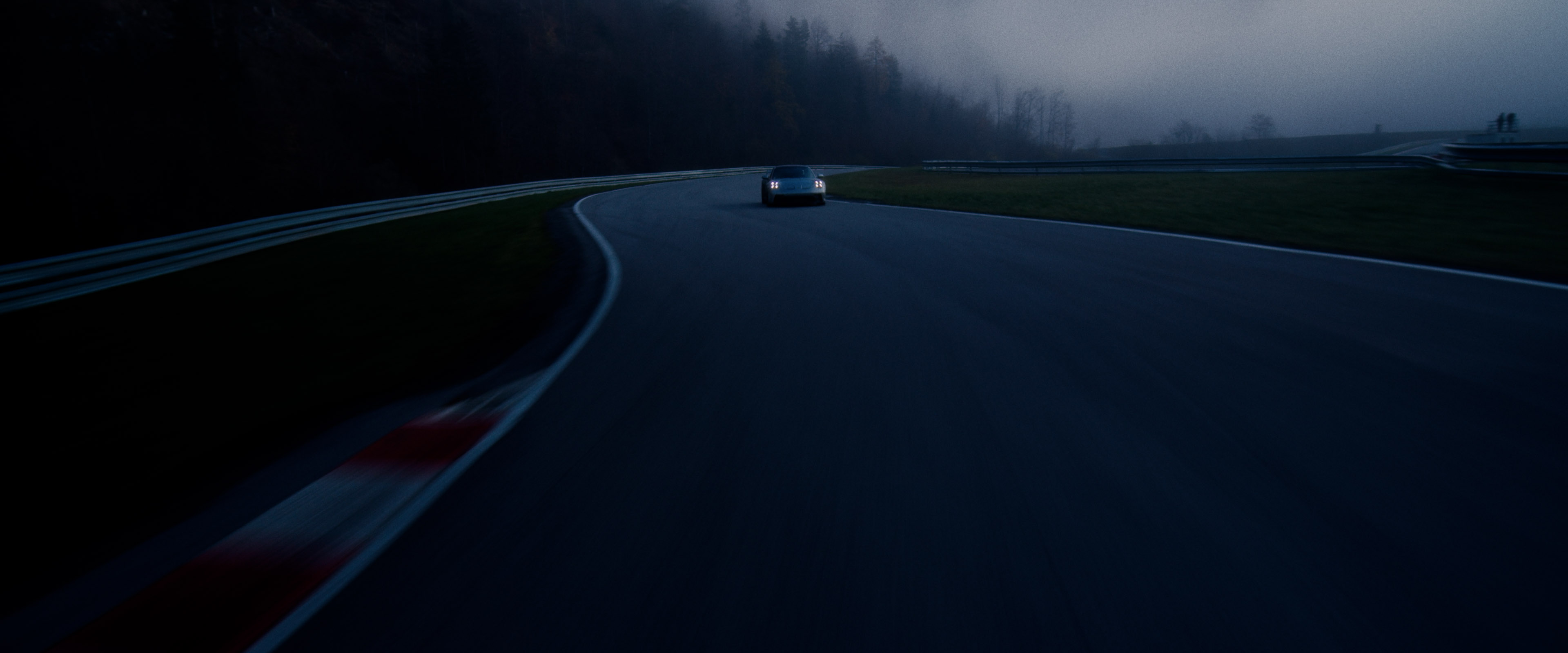 Rear view of a Porsche GT3 entering a foggy racetrack curve at dawn, headlights piercing the mist.