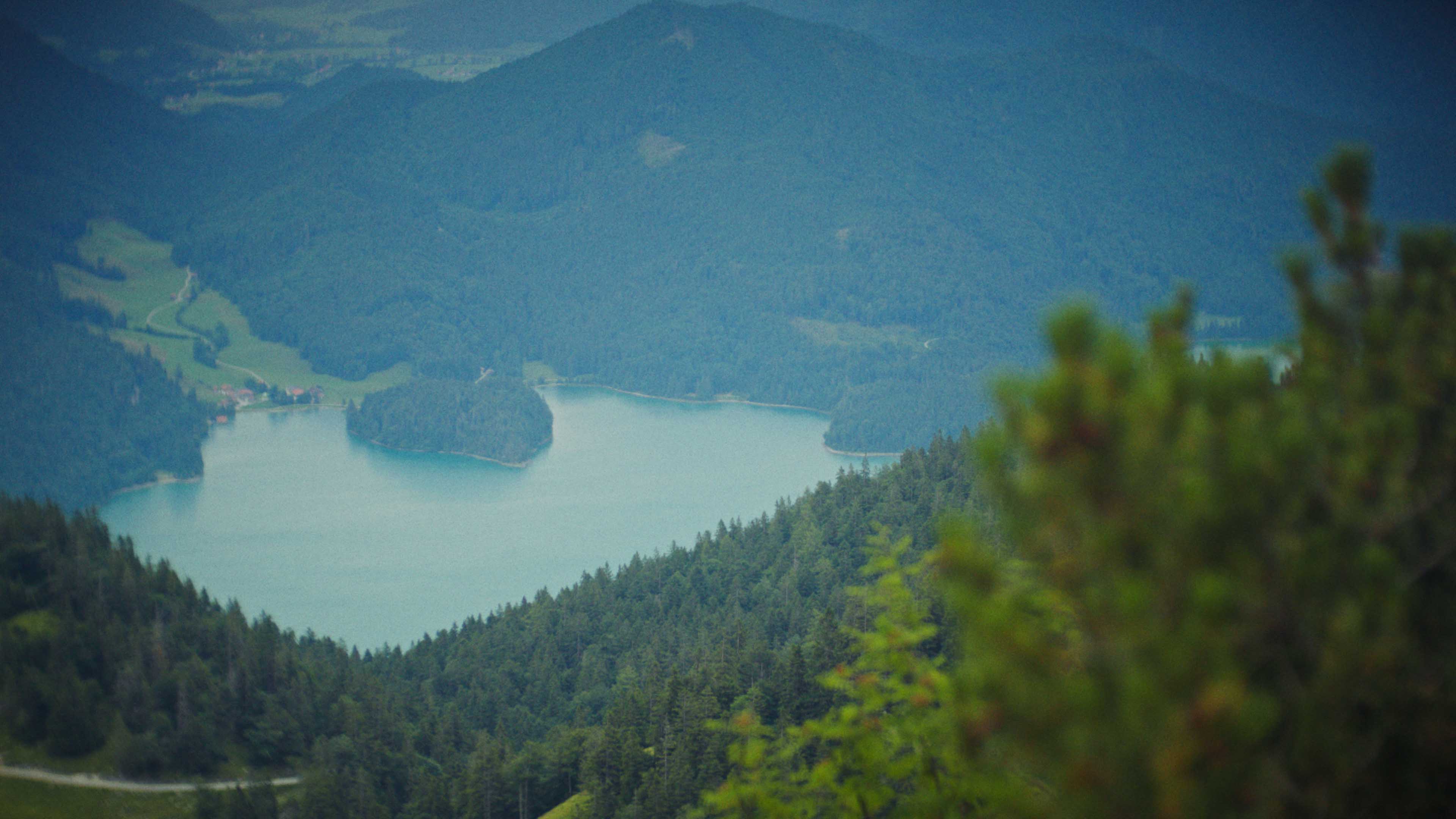 Aerial view of an alpine lakeside road with avalanche gallery; tiny sports car speeds along the shoreline.